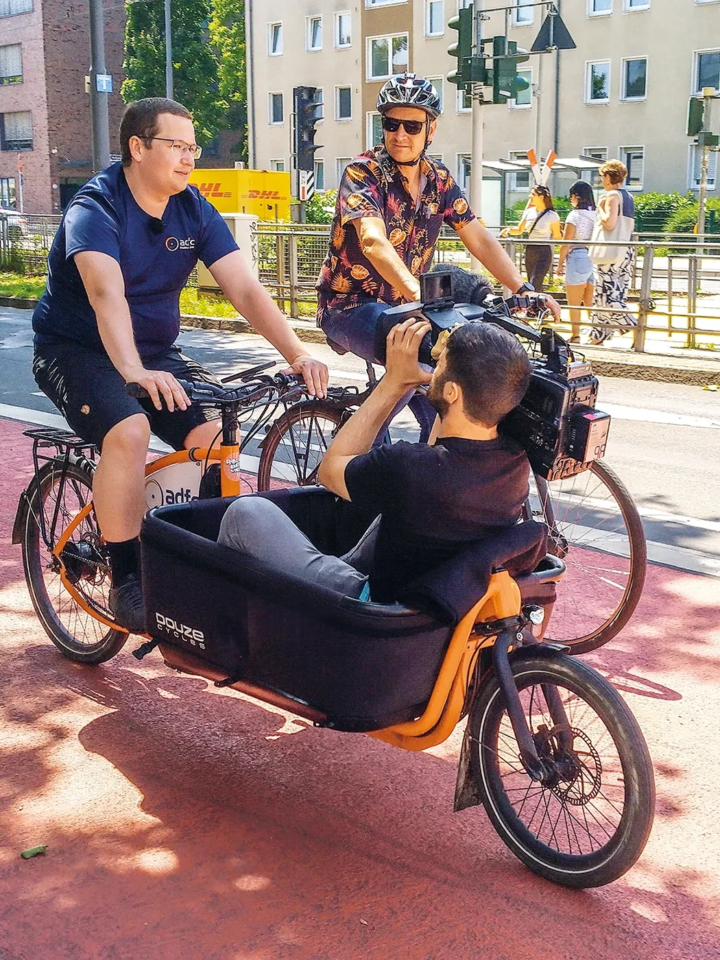 Ein Mann fährt auf dem Lastenrad und wird dabei von einem auf dem Rad liegenden Kameramann gefilmt