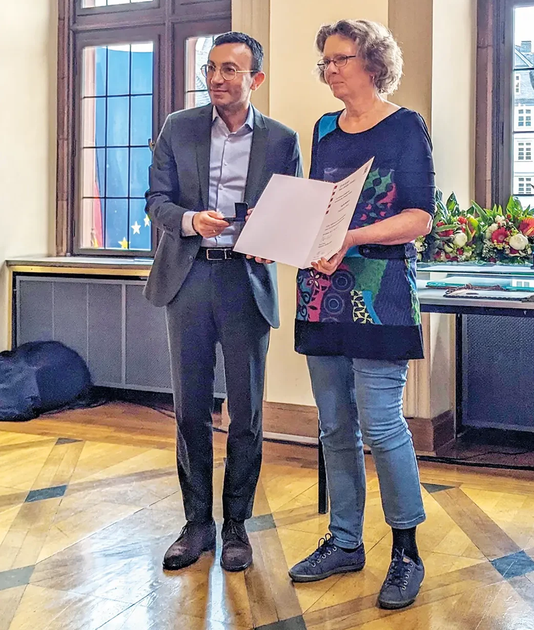 Oberbürgermeister Mike Josef und Sigrid Hubert stehen im Limpurgsaal nebeneinander. Sigrid Hubert hält den Ehrenbrief in der Hand