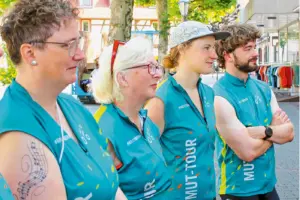 Drei Frauen und ein Mann stehen mit einem T-Shirt mit der Aufschrift MUT-TOUR und blicken auf die Bühne